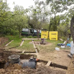 Budowa przydomowej oczyszczalni ścieków z widocznymi elementami instalacji, wykopami, rurami rozsączającymi, studzienką kanalizacyjną, bloczkami Ytong i samochodem dostawczym firmy w tle,...