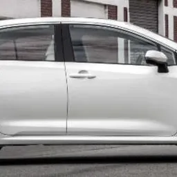 Biała Toyota Corolla Hybrid zaparkowana na ulicy z ceglanymi budynkami w tle, widok z boku.