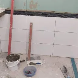 Białe płytki ścienne ułożone do połowy wysokości w trakcie prac wykończeniowych w łazience, widoczne narzędzia glazurnicze, poziomice i wiadro z klejem.