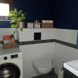 Nowoczesna łazienka z granatową ścianą, białymi płytkami z czarnym paskiem dekoracyjnym, pralką Samsung, wiszącą toaletą i rośliną doniczkową na parapecie.