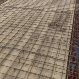 Siatka zbrojeniowa ułożona na drewnianym szalunku, przygotowana do zalania betonem, widoczne ceglane pustaki na obrzeżach, w tle budowlany kontener i trawa.