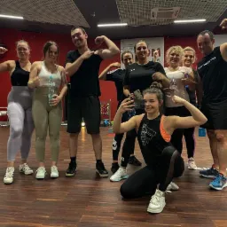Grupa uśmiechniętych osób pozuje, prezentując mięśnie po treningu w sali fitness, jedna osoba robi selfie