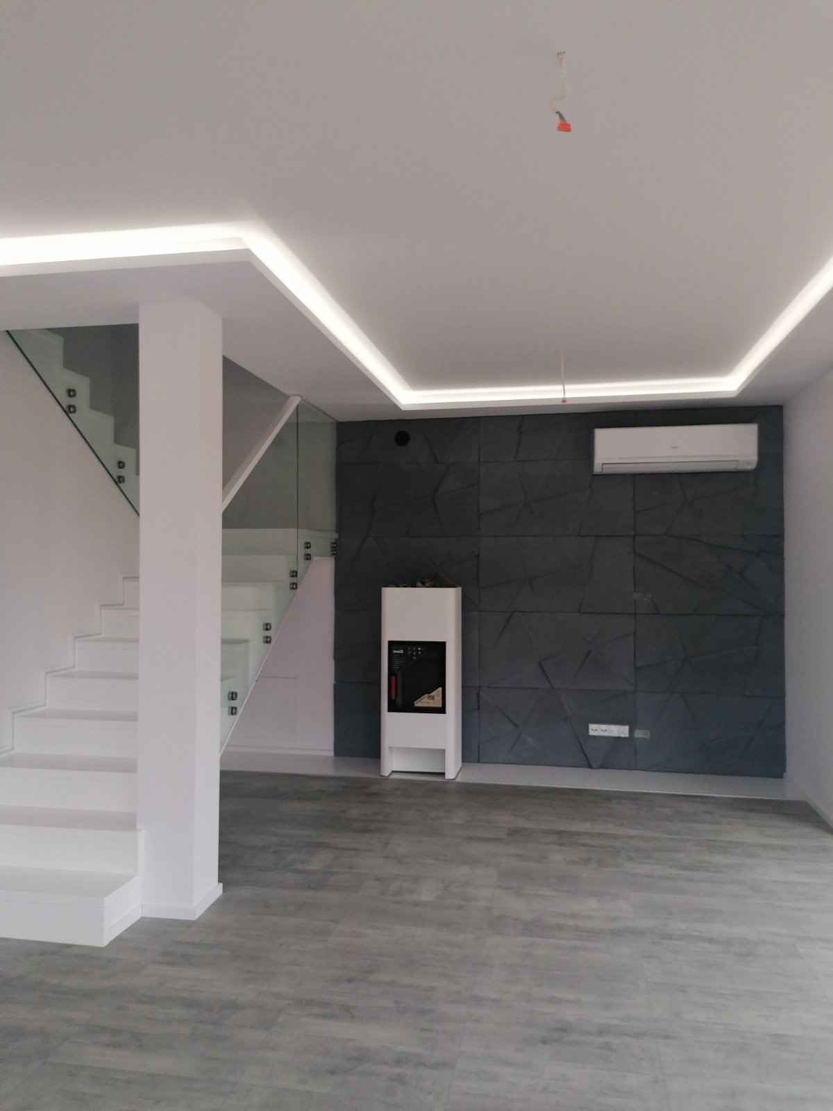 Nowoczesne wnętrze z białymi schodami z szklaną balustradą, szarą ścianą z trójwymiarowych paneli, grzejnikiem elektrycznym, klimatyzacją i oświetleniem LED w suficie podwieszanym.