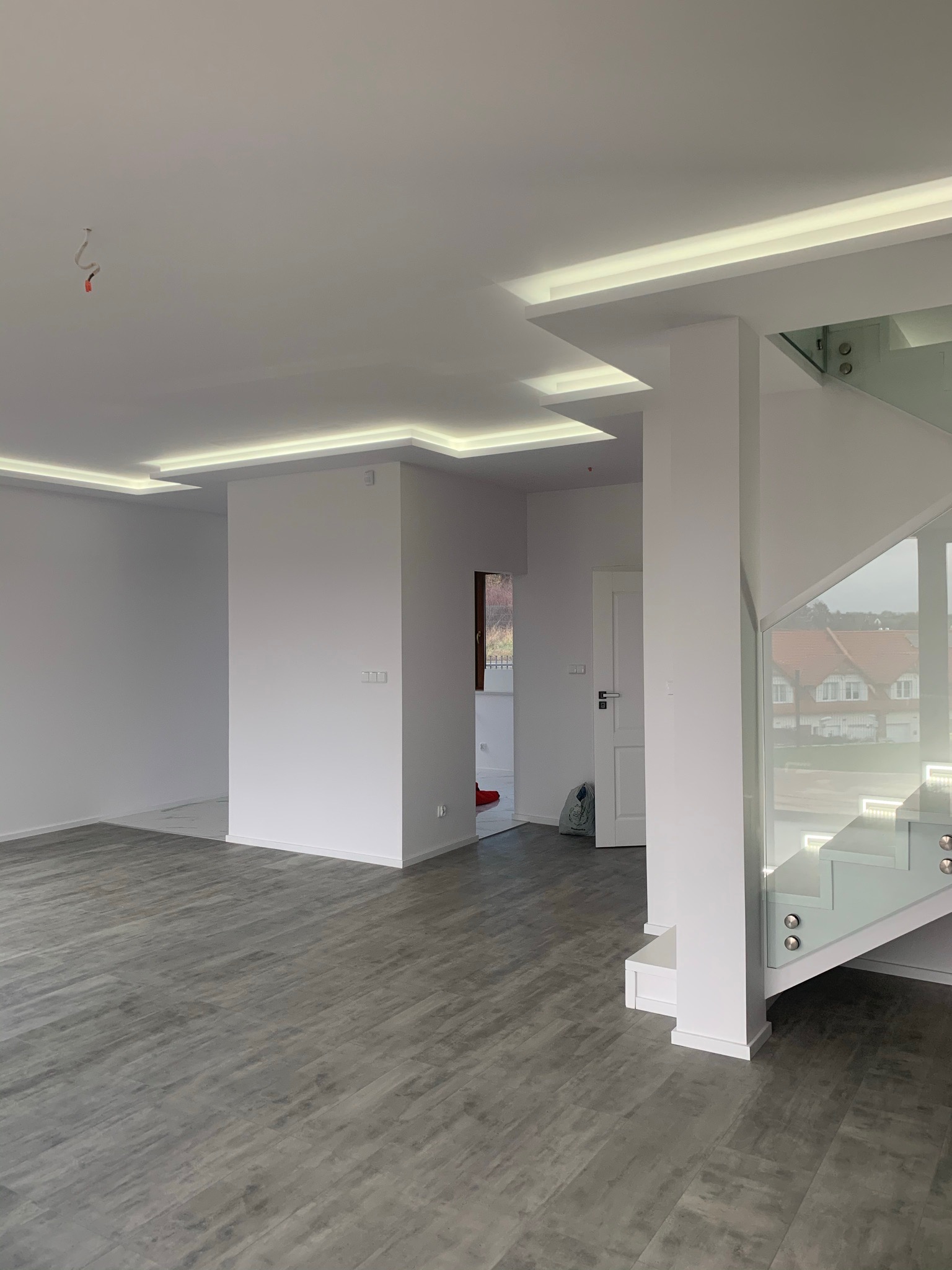 Nowoczesne wnętrze z szarą podłogą imitującą drewno, białymi ścianami, podwieszanym sufitem z oświetleniem LED i szklaną balustradą schodów, widok na dom za oknem.