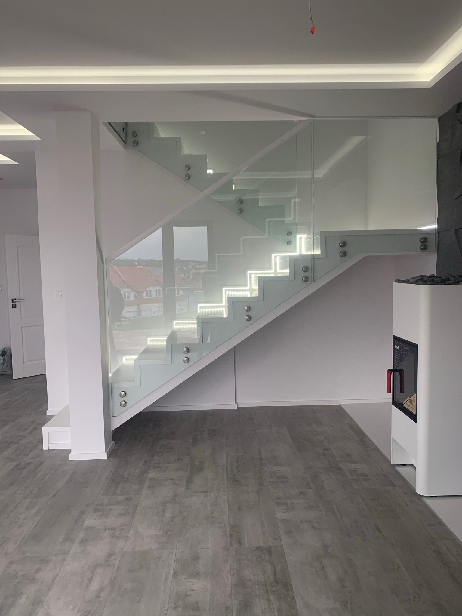 Nowoczesne, białe schody z podświetleniem LED na stopniach i szklanymi balustradami, widoczne z perspektywy podłogi wyłożonej panelami w odcieniach szarości.