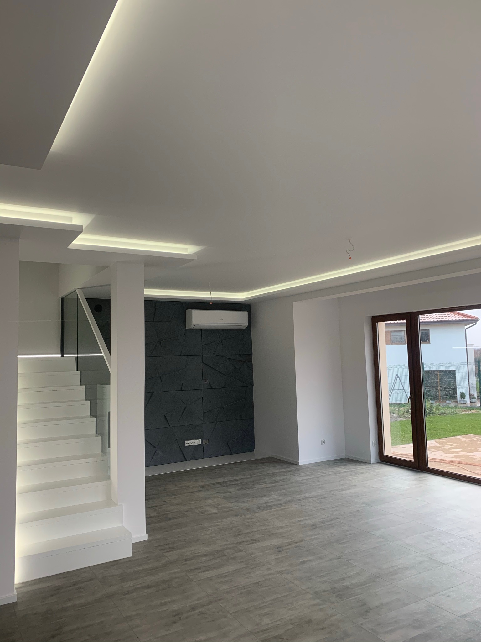 Nowoczesne wnętrze z białymi schodami z podświetleniem LED, szarą ścianą z geometrycznym wzorem, klimatyzatorem i dużym oknem balkonowym z widokiem na ogród.