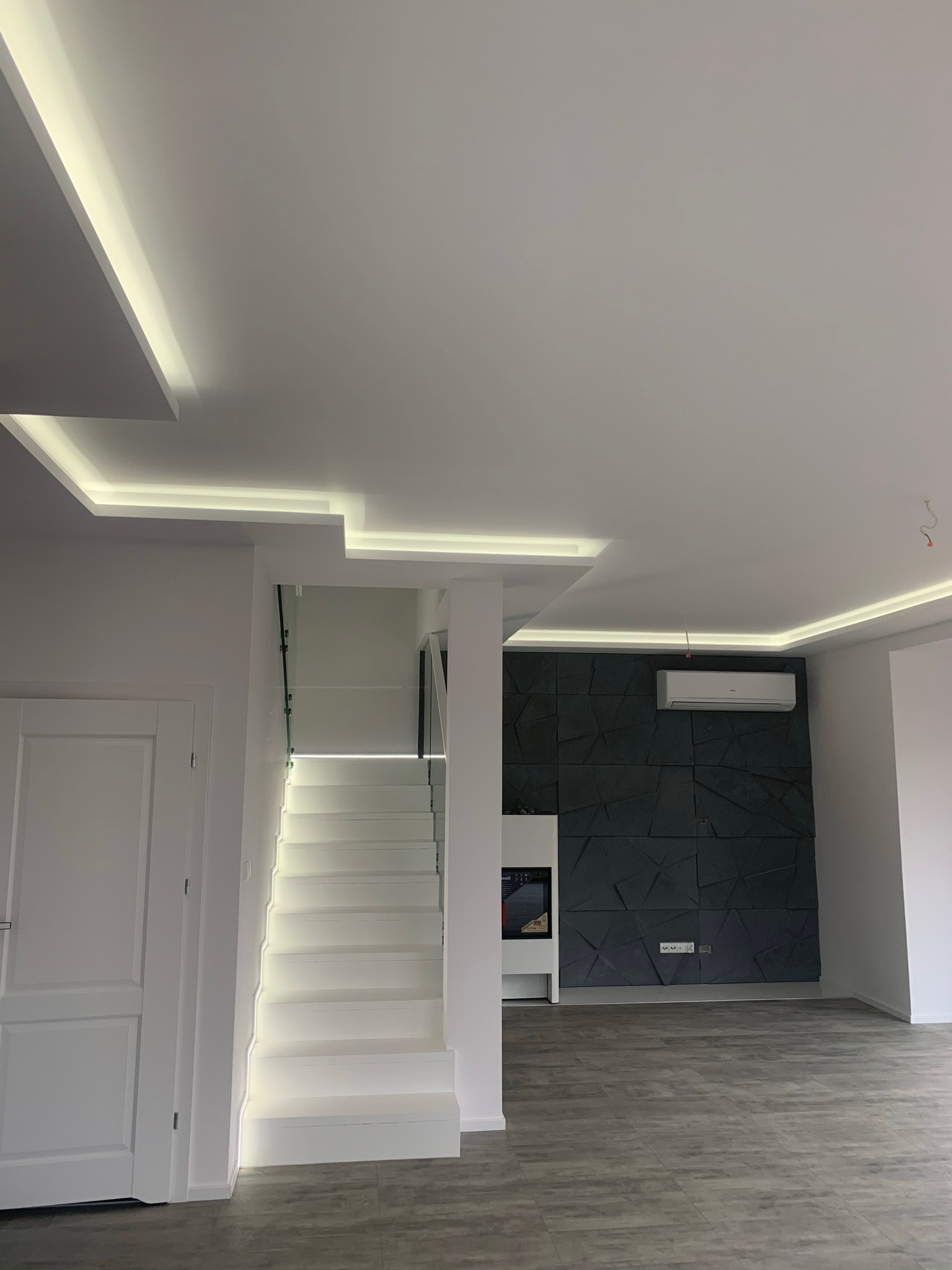 Nowoczesne wnętrze z podświetlonymi schodami, dekoracyjną ścianą z geometrycznym wzorem, klimatyzacją i oświetleniem LED w suficie.