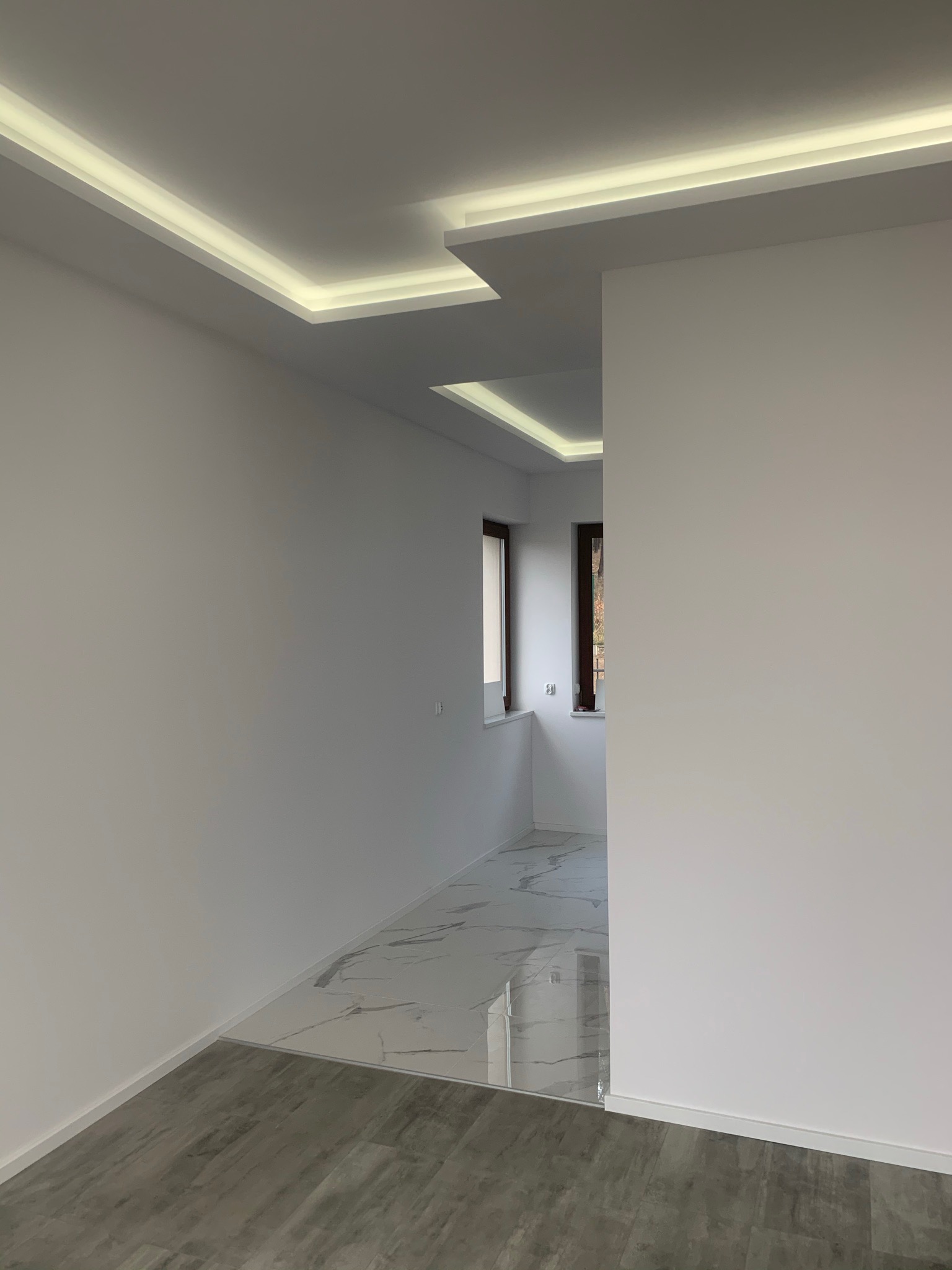 Nowoczesne wnętrze z białymi ścianami, podwieszanym sufitem z oświetleniem LED i dwoma rodzajami podłóg: imitacja drewna i marmurowa posadzka.