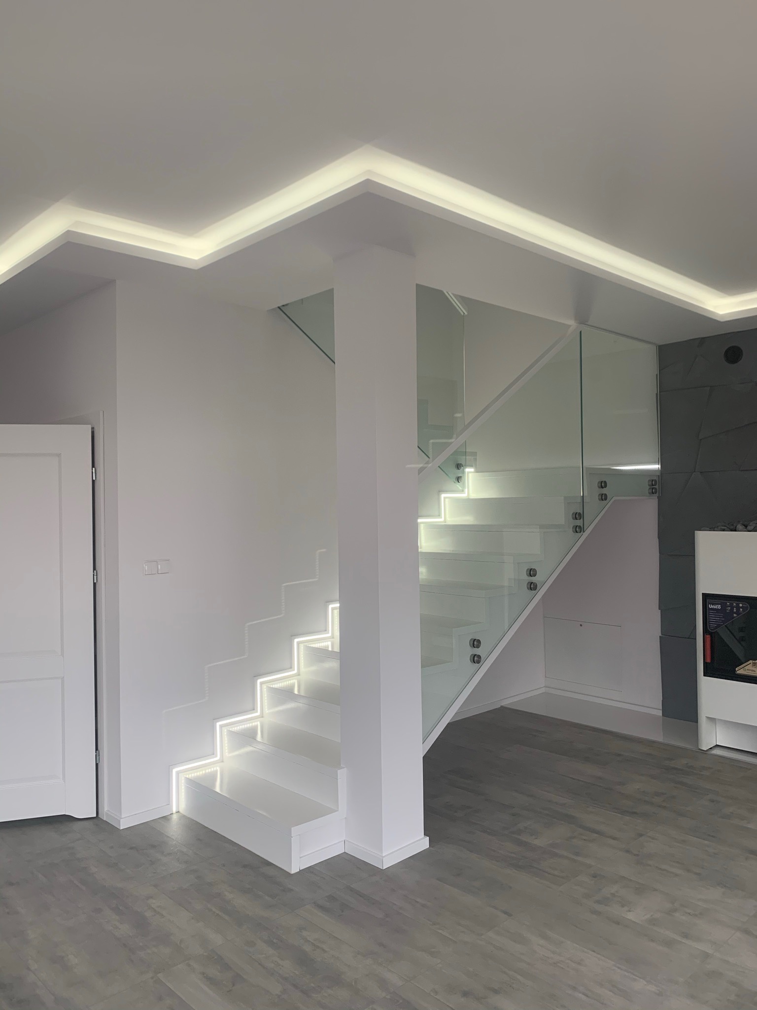 Nowoczesne, białe schody z podświetleniem LED na stopniach i szklaną balustradą, widok z dołu, szare panele podłogowe, białe ściany i drzwi.