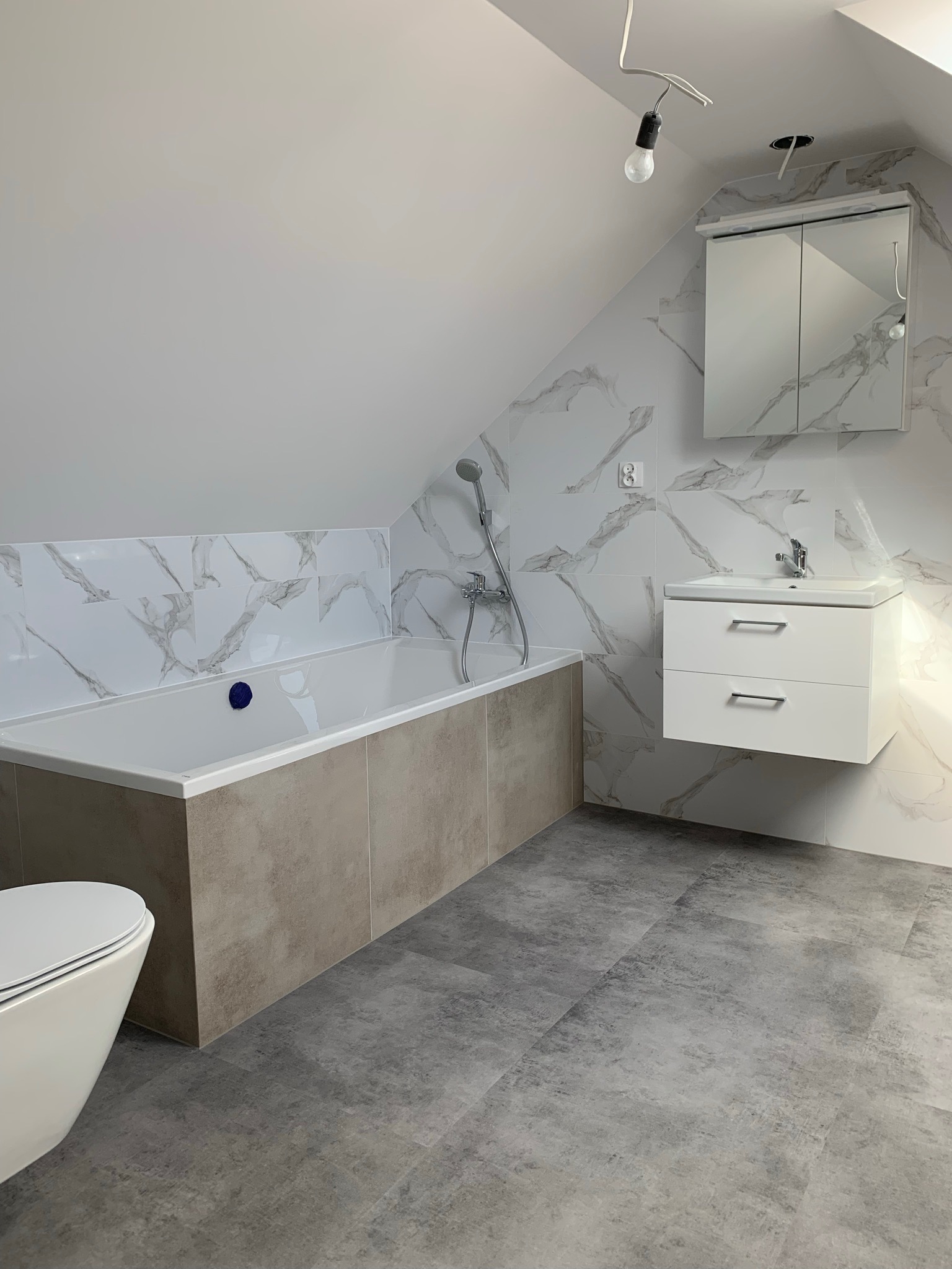 Nowoczesna łazienka na poddaszu z marmurowymi płytkami na ścianach, szarą podłogą, białą armaturą, wiszącą szafką z umywalką i wanną obudowaną płytkami w odcieniach beżu, podwieszona toaleta.