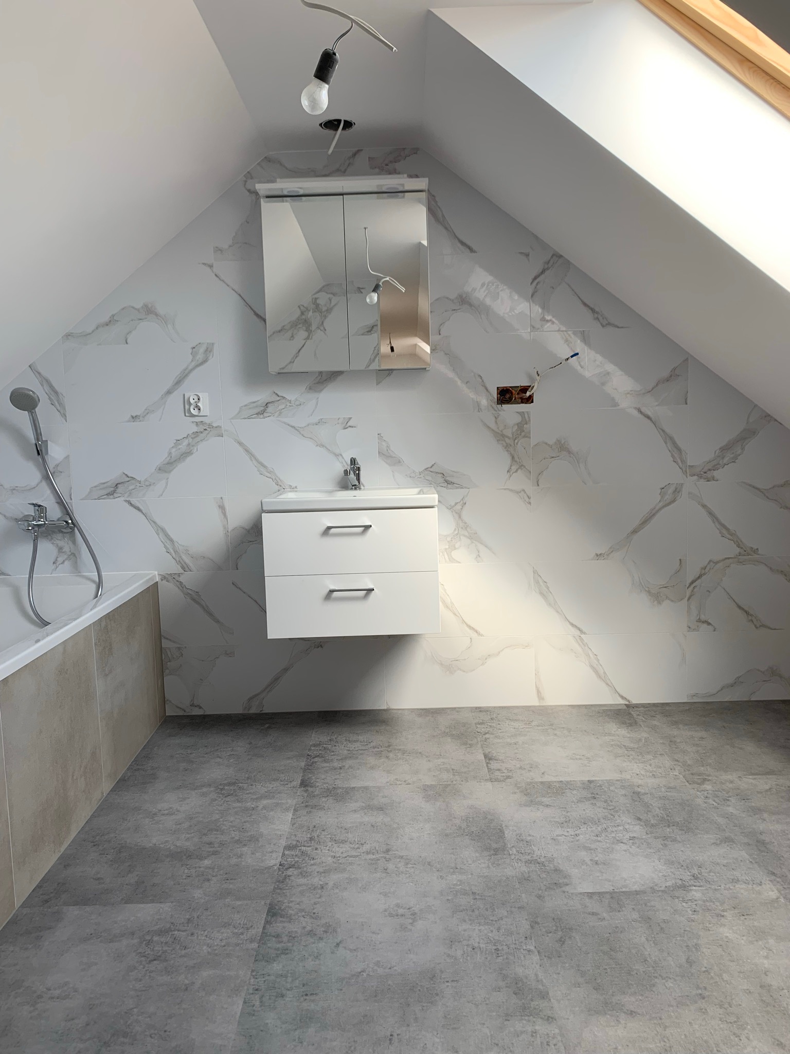 Nowoczesna łazienka na poddaszu z białymi płytkami imitującymi marmur na ścianach, szarą podłogą, białą szafką z umywalką i lustrem, widoczne niedokończone podłączenie elektryczne