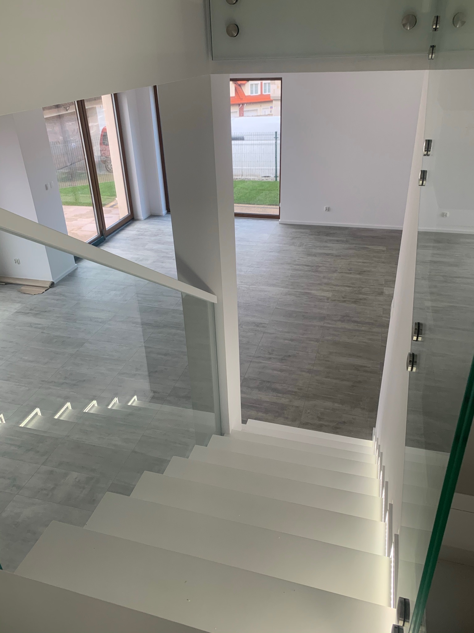 Nowoczesne wnętrze z białymi schodami z podświetleniem LED, szklane balustrady, szare płytki podłogowe imitujące drewno, duże okna z widokiem na ogród.