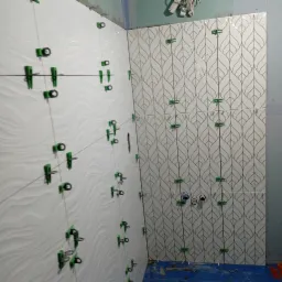 Układanie białych płytek z falistym wzorem i płytek z motywem liści na ścianie w łazience z użyciem klipsów poziomujących.