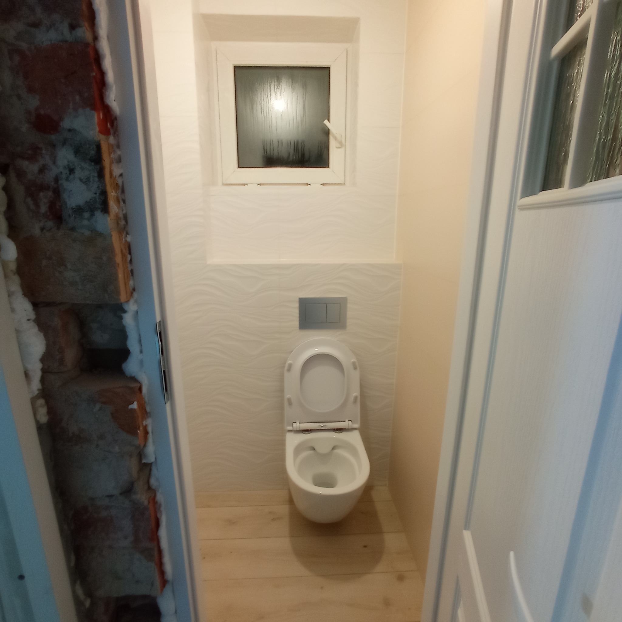 Nowoczesna toaleta wisząca z otwartą klapą w wyremontowanej łazience, widok przez częściowo zamontowane drzwi, odsłonięta cegła i pianka montażowa w ościeżnicy.