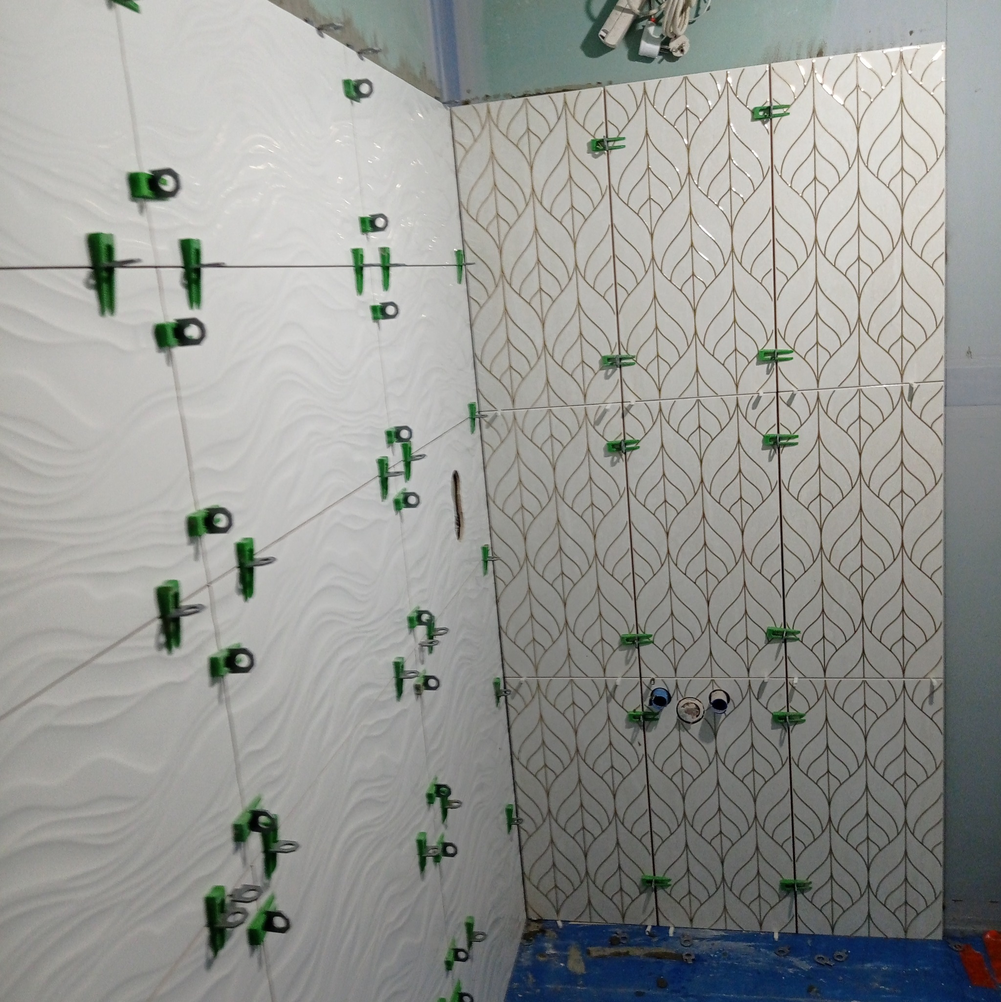 Układanie białych płytek z falistym wzorem i płytek z motywem liści na ścianie w łazience z użyciem klipsów poziomujących.