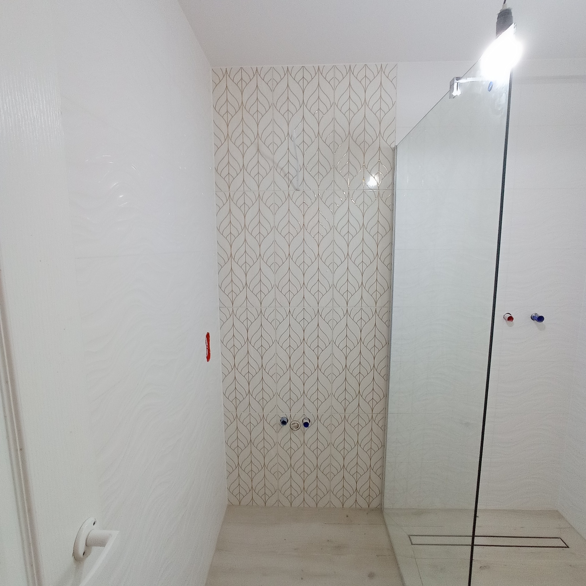 Wykończona łazienka z kabiną prysznicową typu walk-in, białe płytki z delikatnym wzorem liści na jednej ścianie, widoczne przyłącza wodne i odpływ liniowy.