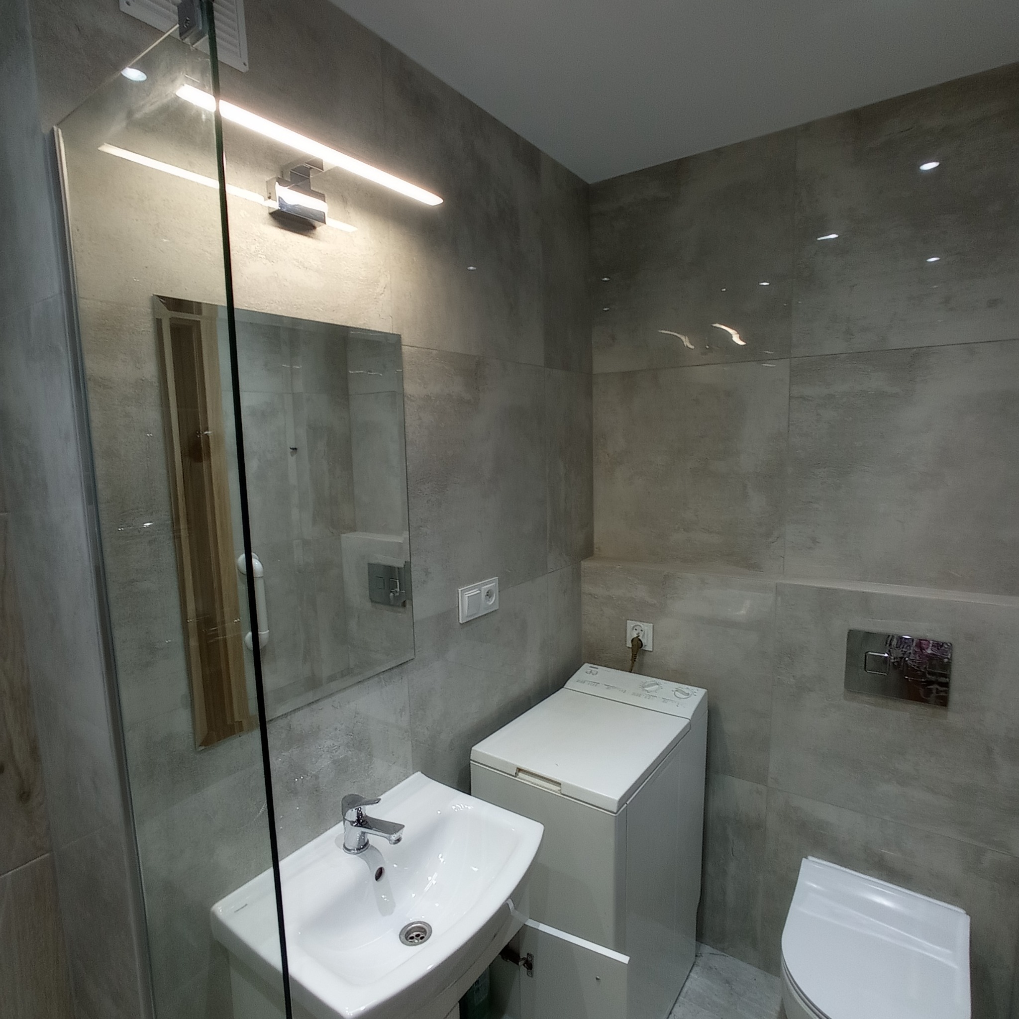 Nowoczesna łazienka w odcieniach szarości z płytkami imitującymi beton, umywalką z szafką, pralką i toaletą podwieszaną. Oświetlenie LED nad lustrem.