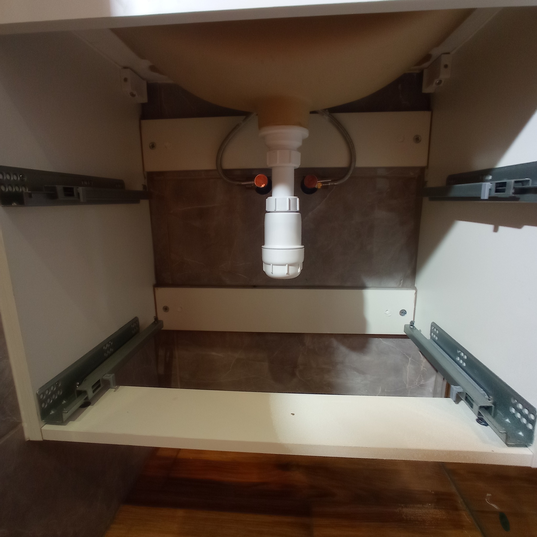 Wnętrze szafki umywalkowej z widocznym syfonem, instalacją wodną i prowadnicami szuflad, marmurowe płytki na ścianie.