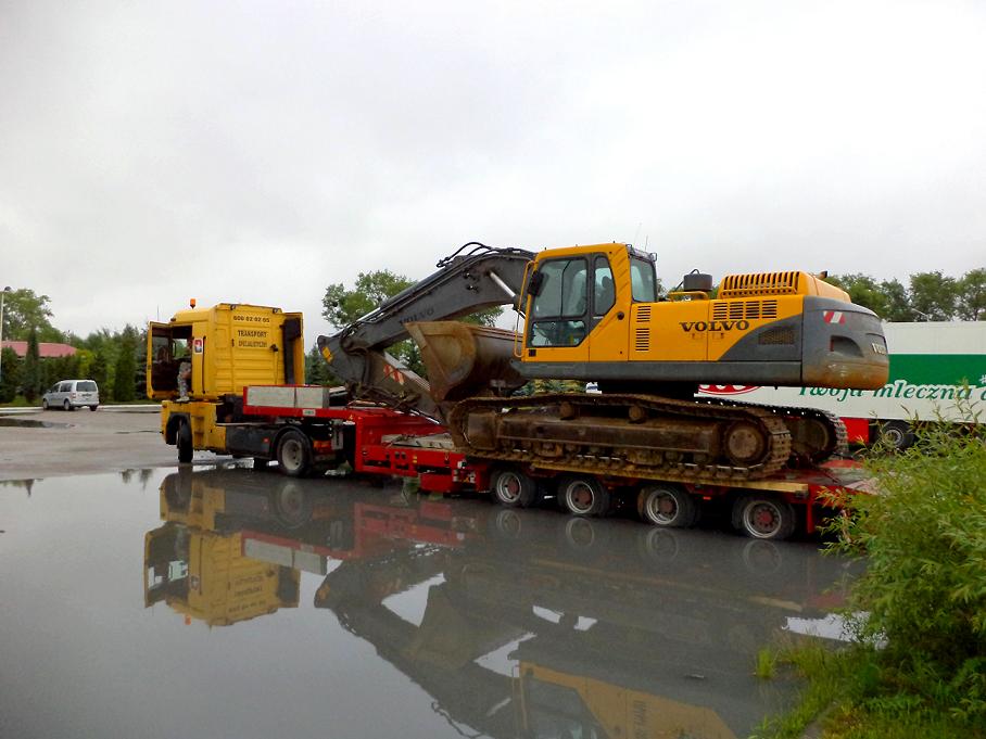 Żółta koparka Volvo na niskopodwoziowej platformie transportowej, odbijająca się w kałuży na placu manewrowym, z żółtą kabiną ciągnika siodłowego w tle.