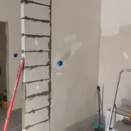 Wnętrze w trakcie prac remontowych: ściana z bloków, częściowo otynkowana ściana z widocznymi puszkami elektrycznymi, poziomica, wiadro z zaprawą Atlas i drabina aluminiowa.