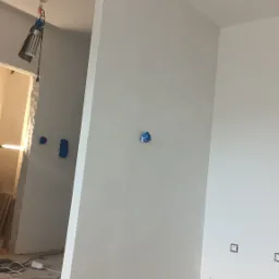 Świeżo pomalowane na biało ściany w trakcie remontu, zabezpieczone gniazdka elektryczne niebieską taśmą malarską, podłoga zabezpieczona kartonami, widoczna lampa budowlana.