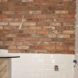 Ściana łazienki w trakcie wykończenia: cegła dekoracyjna u góry, białe, prostokątne płytki poniżej, widoczne wyprowadzenia instalacji wodnej i elektrycznej.