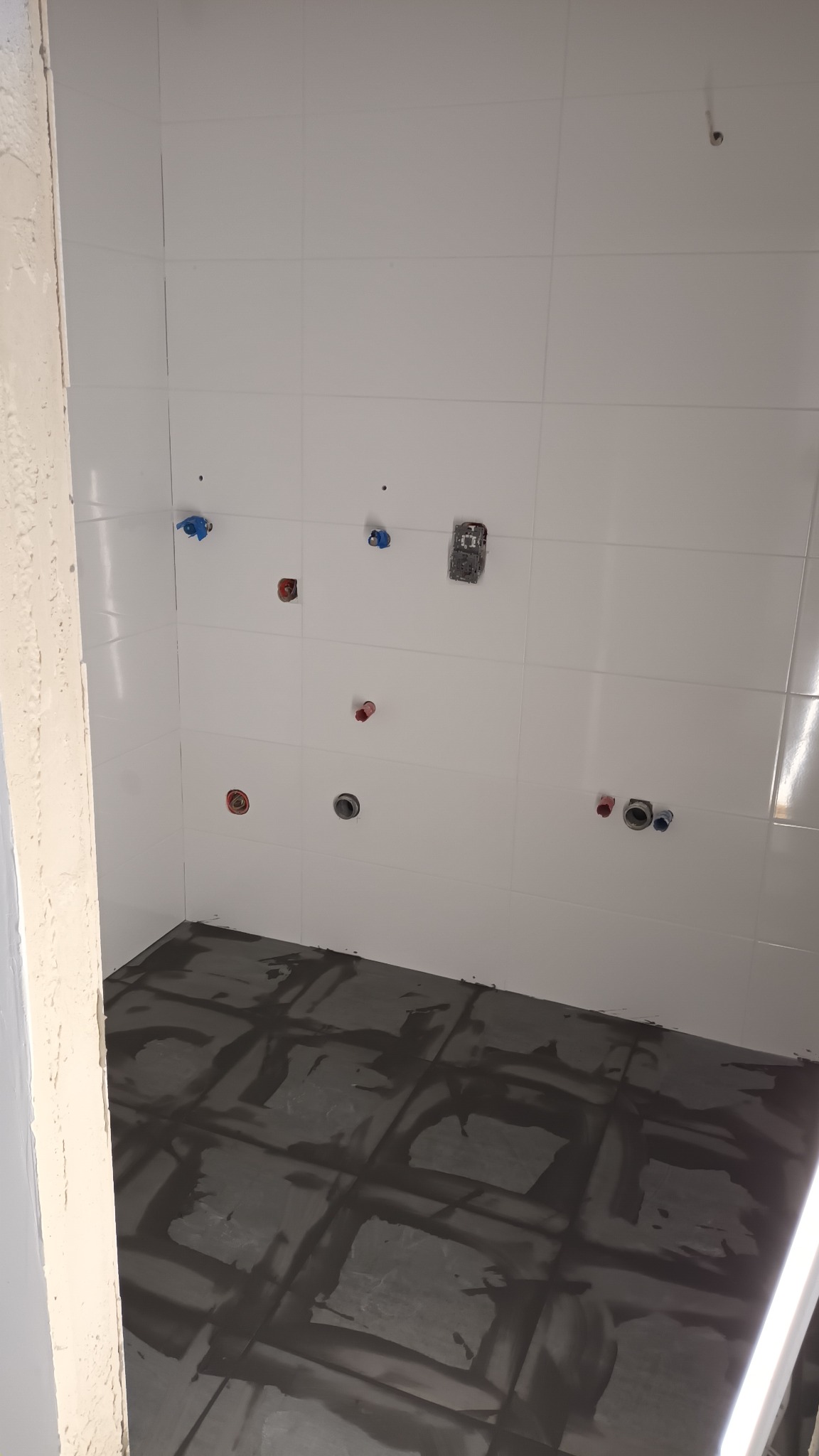 Przygotowana do fugowania łazienka z białymi płytkami ściennymi i ciemną, zafugowaną podłogą. Widoczne wystające rury instalacji wodnej i elektrycznej.