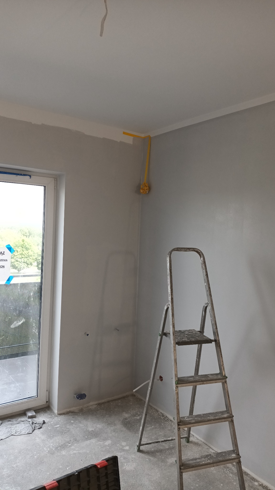 Wnętrze w trakcie prac wykończeniowych: świeżo pomalowane szare ściany, aluminiowa drabina oparta o ścianę, widoczna instalacja elektryczna zabezpieczona żółtą taśmą, okno balkonowe z widokiem...