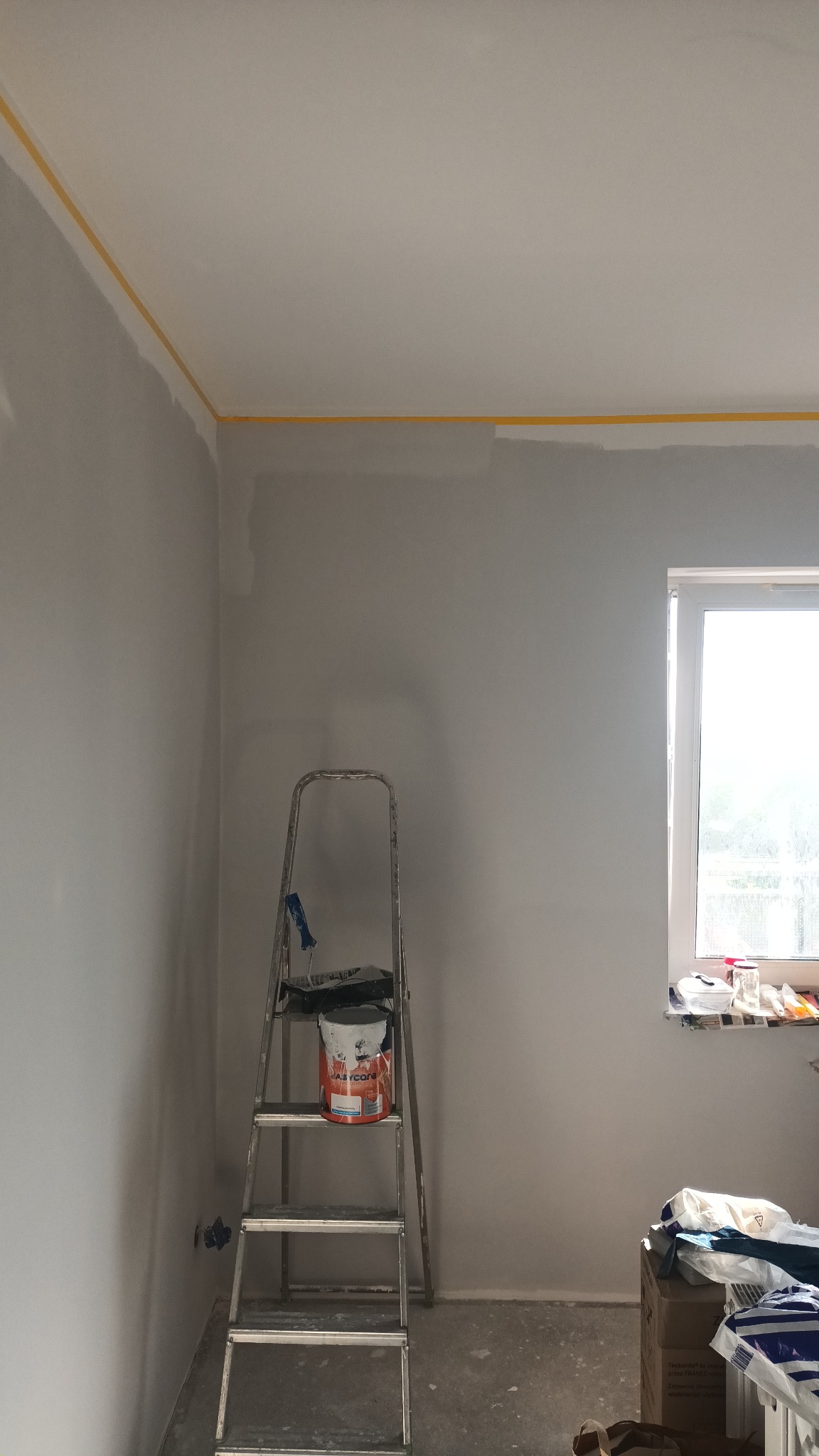 Pokój w trakcie malowania: szara ściana z żółtą taśmą malarską przy suficie, drabina z wiadrem farby Easycare, częściowo pomalowana ściana, okno w tle.