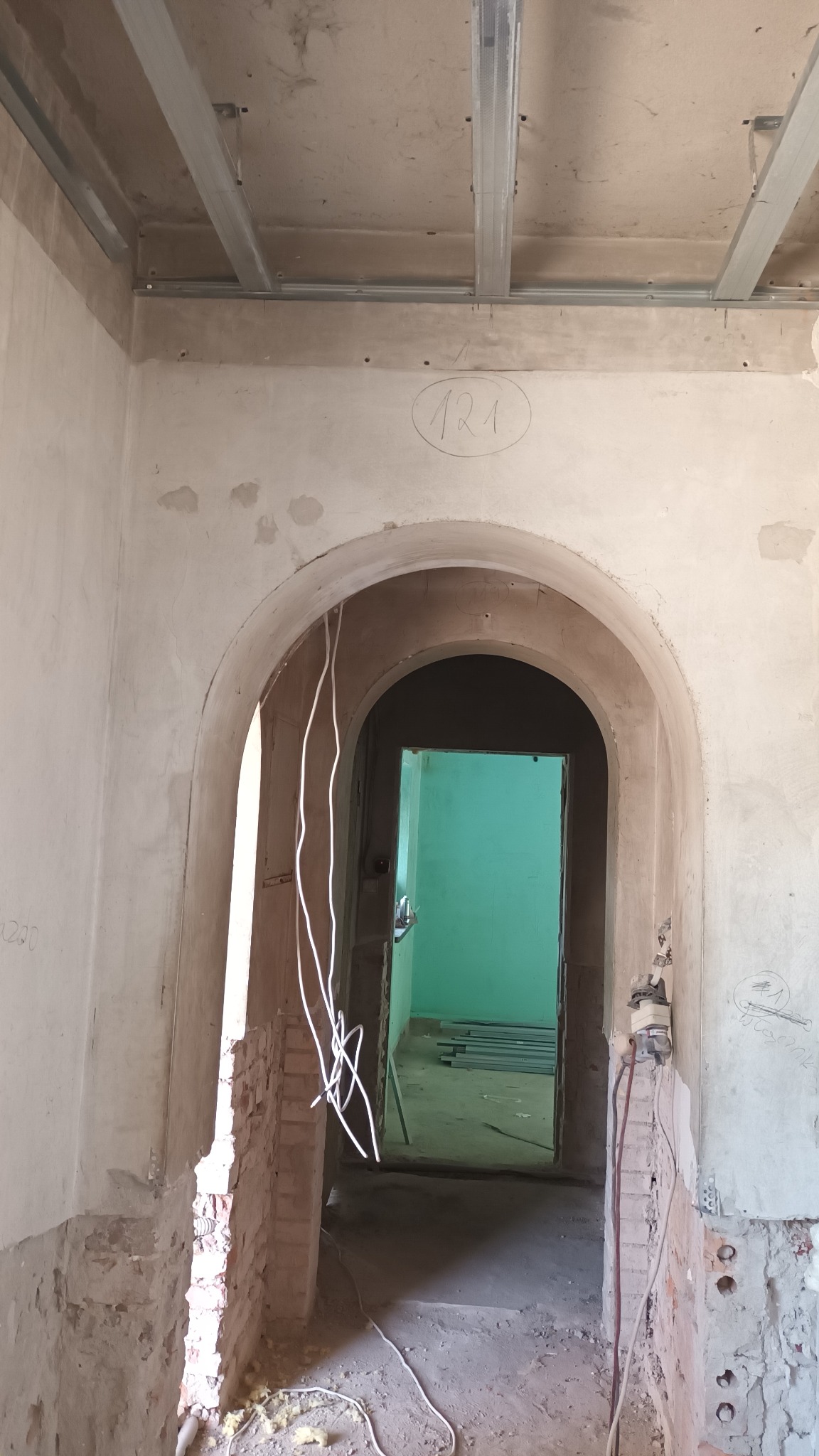 Wnętrze w trakcie remontu: widoczne surowe ściany z odsłoniętą cegłą, łukowate przejście do pomieszczenia z zielonymi ścianami, metalowa konstrukcja sufitu podwieszanego i wiszące kable elektryczne.