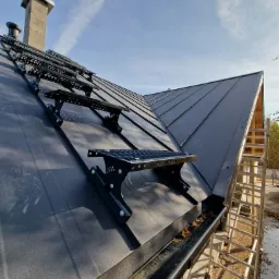 Nowy, grafitowy dach z blachy na rąbek stojący z zamontowanymi stopniami kominiarskimi, widoczne rynny z liśćmi i drabina oparta o ścianę budynku.