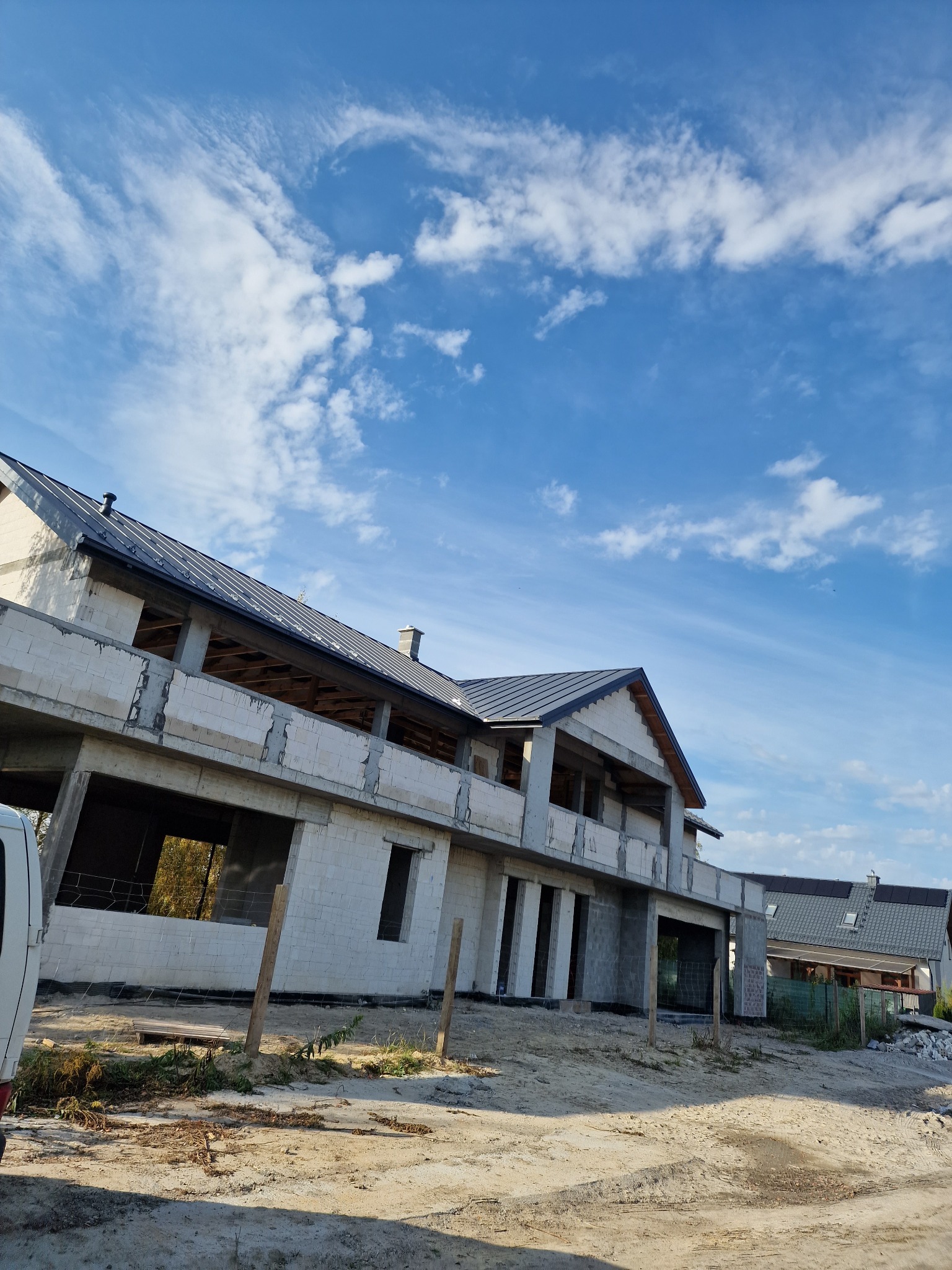 Niedokończony budynek z szarym dachem pod błękitnym niebem z chmurami, widoczne drewniane elementy konstrukcyjne i pustaki.