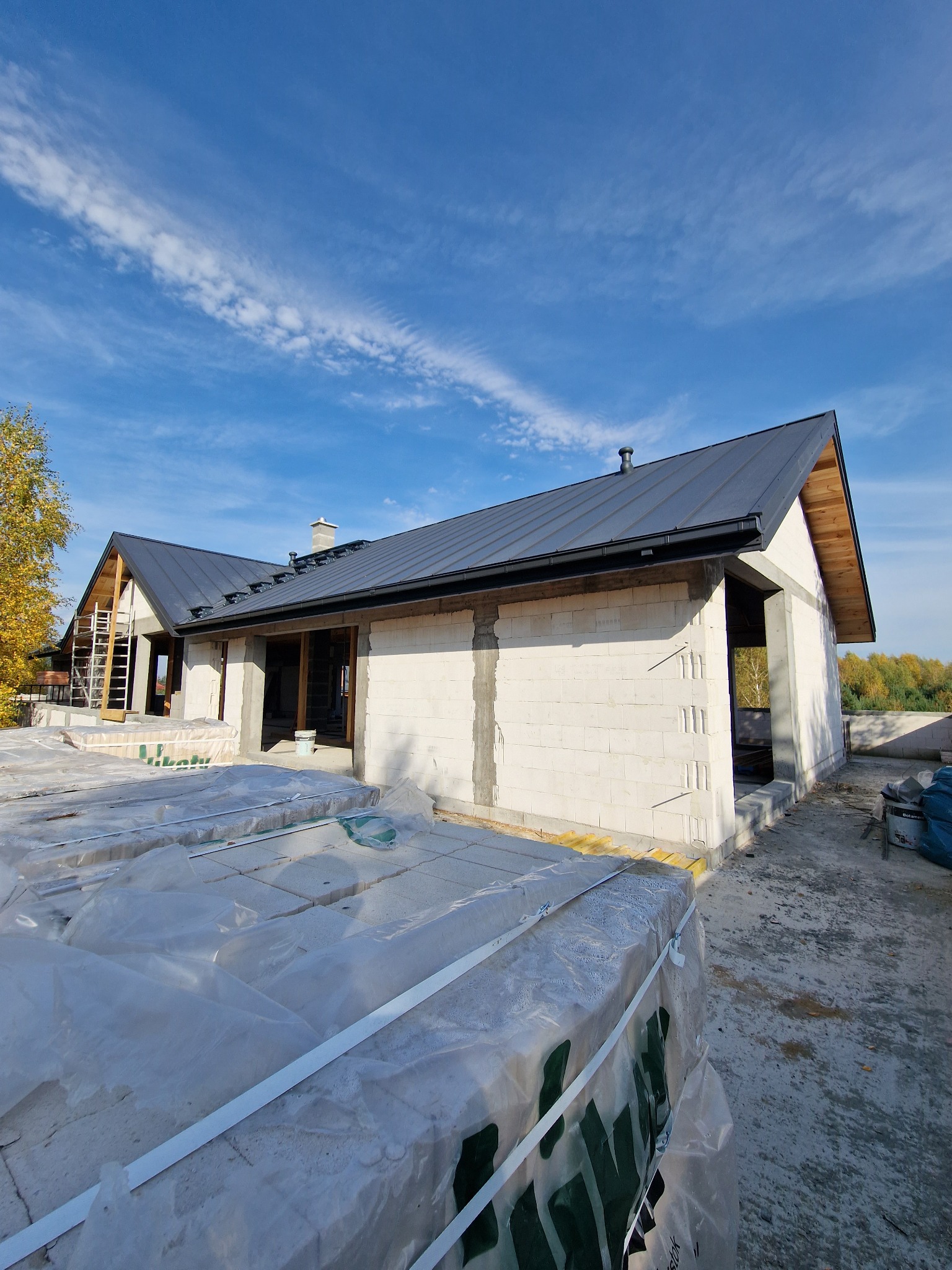 Dom w trakcie budowy z czarnym dachem z blachy na tle błękitnego nieba z lekkimi chmurami, widoczne elementy konstrukcyjne i materiały budowlane.