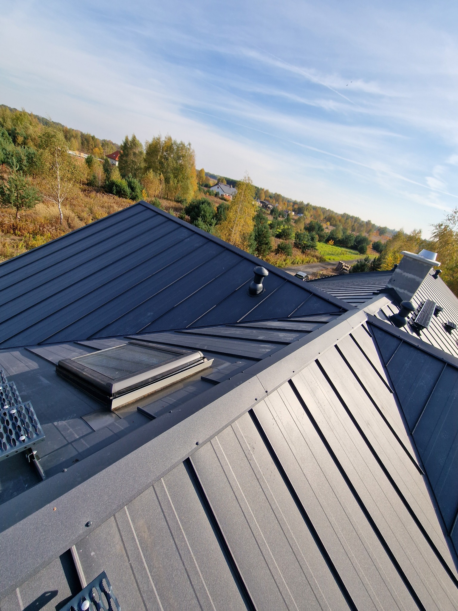 Widok z góry na nowy, ciemnoszary dach panelowy z oknem dachowym, kominem i wentylacją, na tle jesiennego krajobrazu z drzewami i błękitnym niebem.
