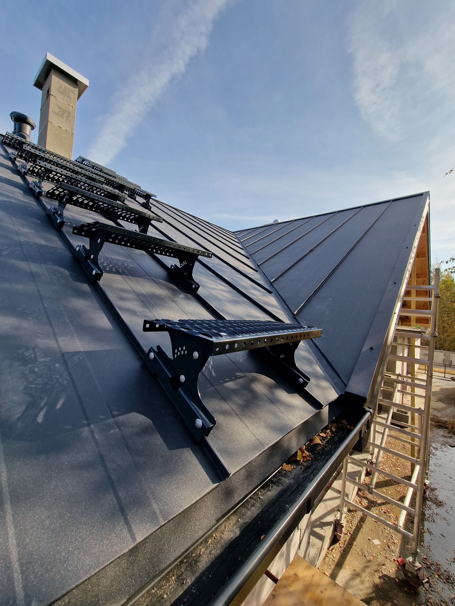 Nowy, grafitowy dach z blachy na rąbek stojący z zamontowanymi stopniami kominiarskimi, widoczne rynny z liśćmi i drabina oparta o ścianę budynku.