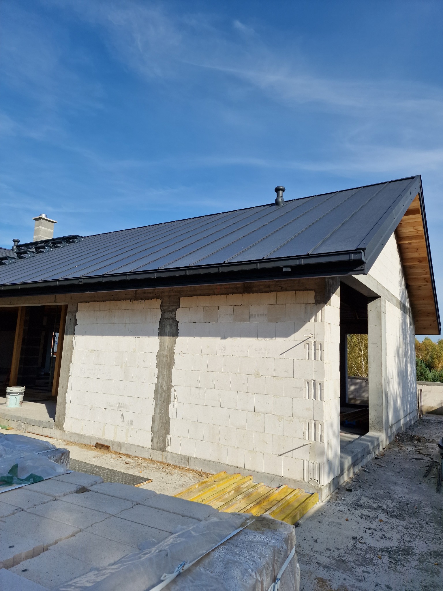 Surowy stan domu w budowie z zamontowanym ciemnoszarym dachem z blachy trapezowej i czarnymi rynnami; widoczne bloczki betonowe, otwory okienne i drzwiowe oraz elementy konstrukcji drewnianej dachu.