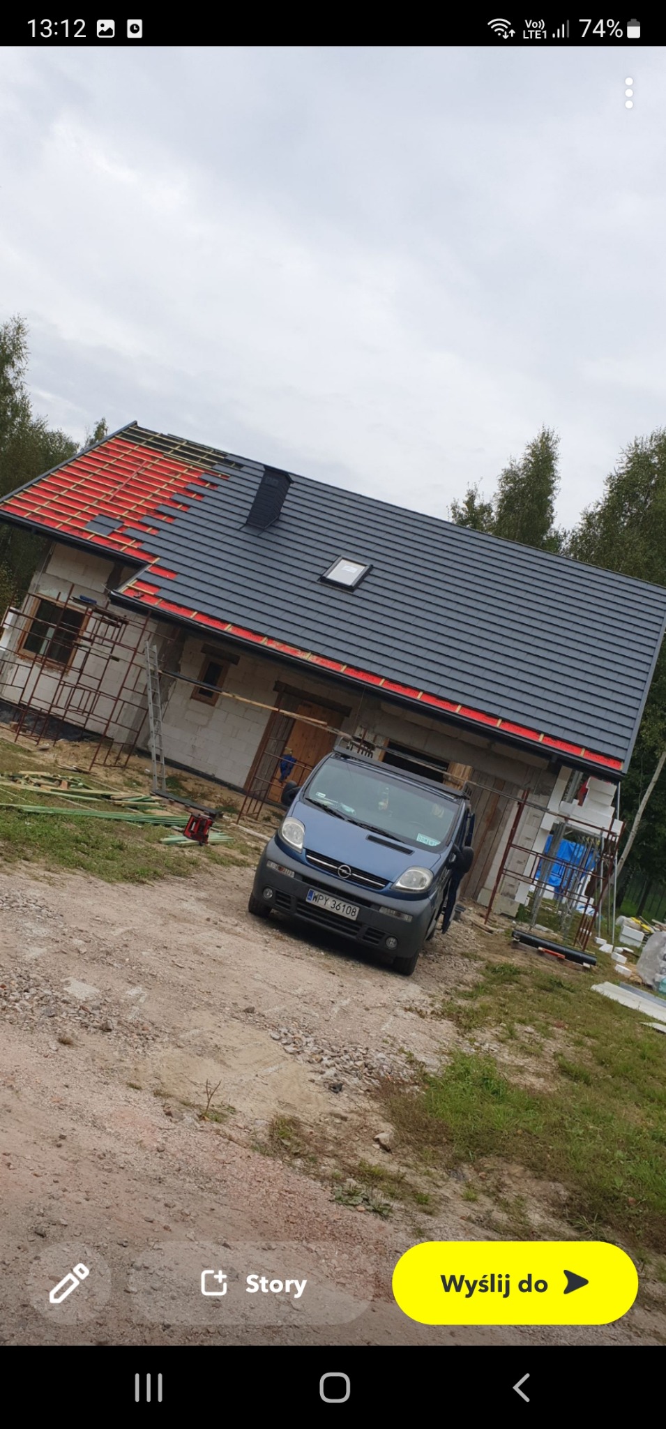 Dom w trakcie budowy z częściowo położonym dachem z grafitowej blachodachówki z oknem dachowym i czerwonymi elementami pod spodem, na pierwszym planie niebieski samochód dostawczy.