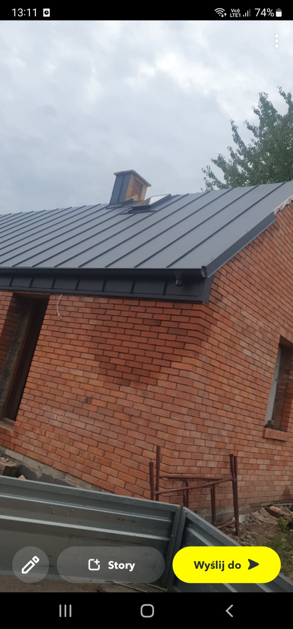 Nowy, grafitowy dach z blachy na domu z czerwonej cegły; widoczne rynny i komin, brak okien; w tle drzewo i pochmurne niebo.