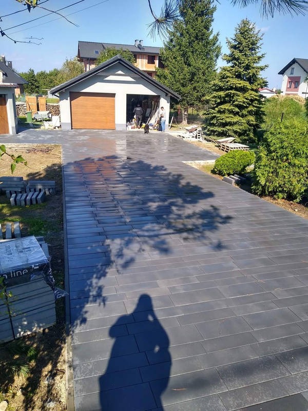Ułożona, grafitowa kostka brukowa na podjeździe do garażu w słoneczny dzień. W tle domy i zieleń. Widoczny cień osoby robiącej zdjęcie i palety z kostką.