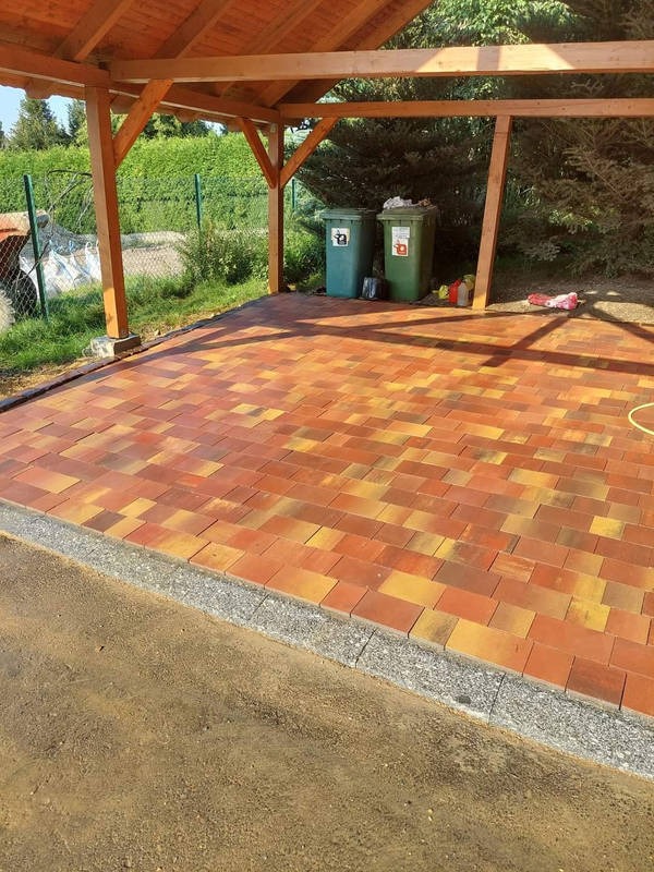 Kolorowa kostka brukowa pod drewnianą wiatą, obrzeża z szarego kamienia. W tle zielone pojemniki na śmieci i ogrodzenie z siatki.