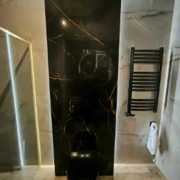 Nowoczesna łazienka z czarną miską WC i płytkami imitującymi marmur z złotymi żyłami, podświetlona listwą LED, z grzejnikiem drabinkowym.