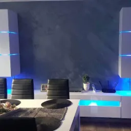 Salon z dekoracyjną, szarą ścianą, podświetlanymi na niebiesko szafkami, czarnymi krzesłami i białym stołem z dekoracjami.