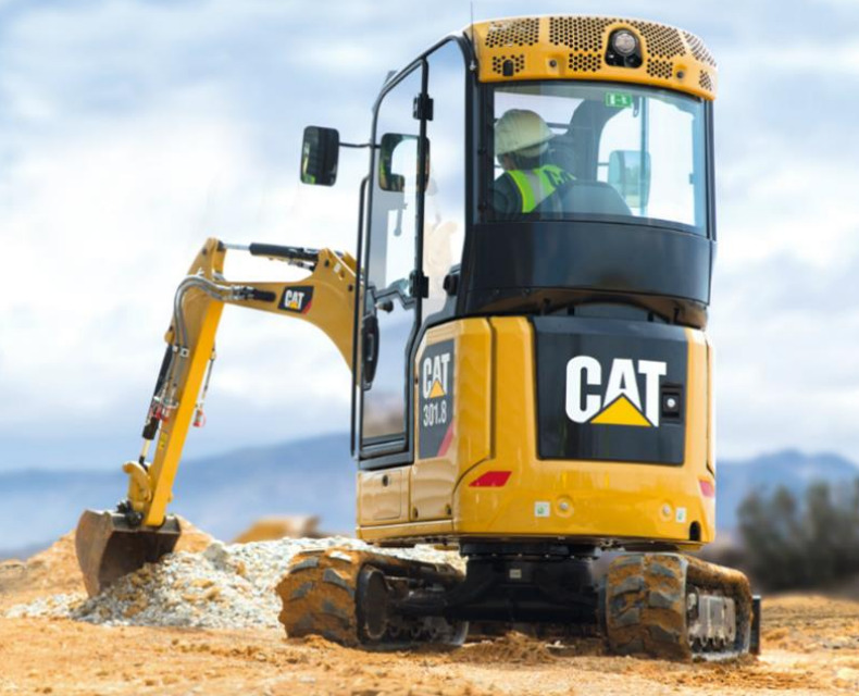 Żółta maszyna budowlana CAT 301.8 z operatorem w kasku i kamizelce odblaskowej, pracująca na placu budowy z górą piasku i żwiru w tle.