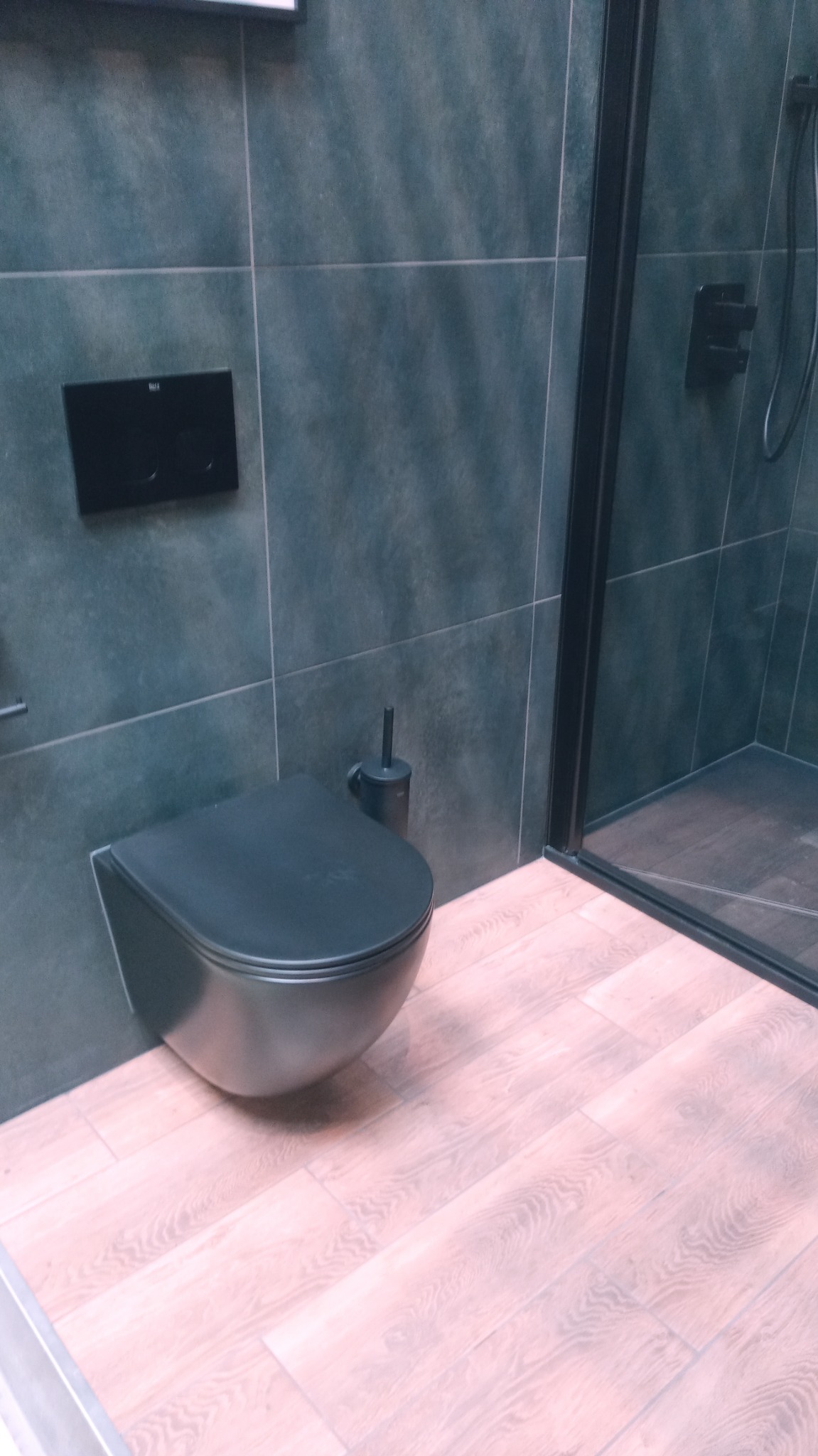 Nowoczesna łazienka z podwieszaną toaletą w kolorze antracytu, ciemnoszare płytki na ścianach i podłoga imitująca drewno w odcieniach różu i beżu, kabina prysznicowa z czarnymi profilami.