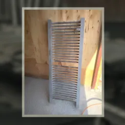 Metalowy grzejnik łazienkowy po piaskowaniu, widoczne ślady obróbki, surowe drewno w tle.