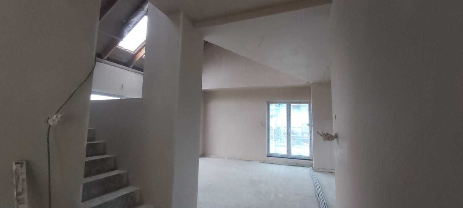 Surowe wnętrze domu w trakcie wykańczania, świeżo położone gładzie na ścianach, widoczne schody i okno dachowe.