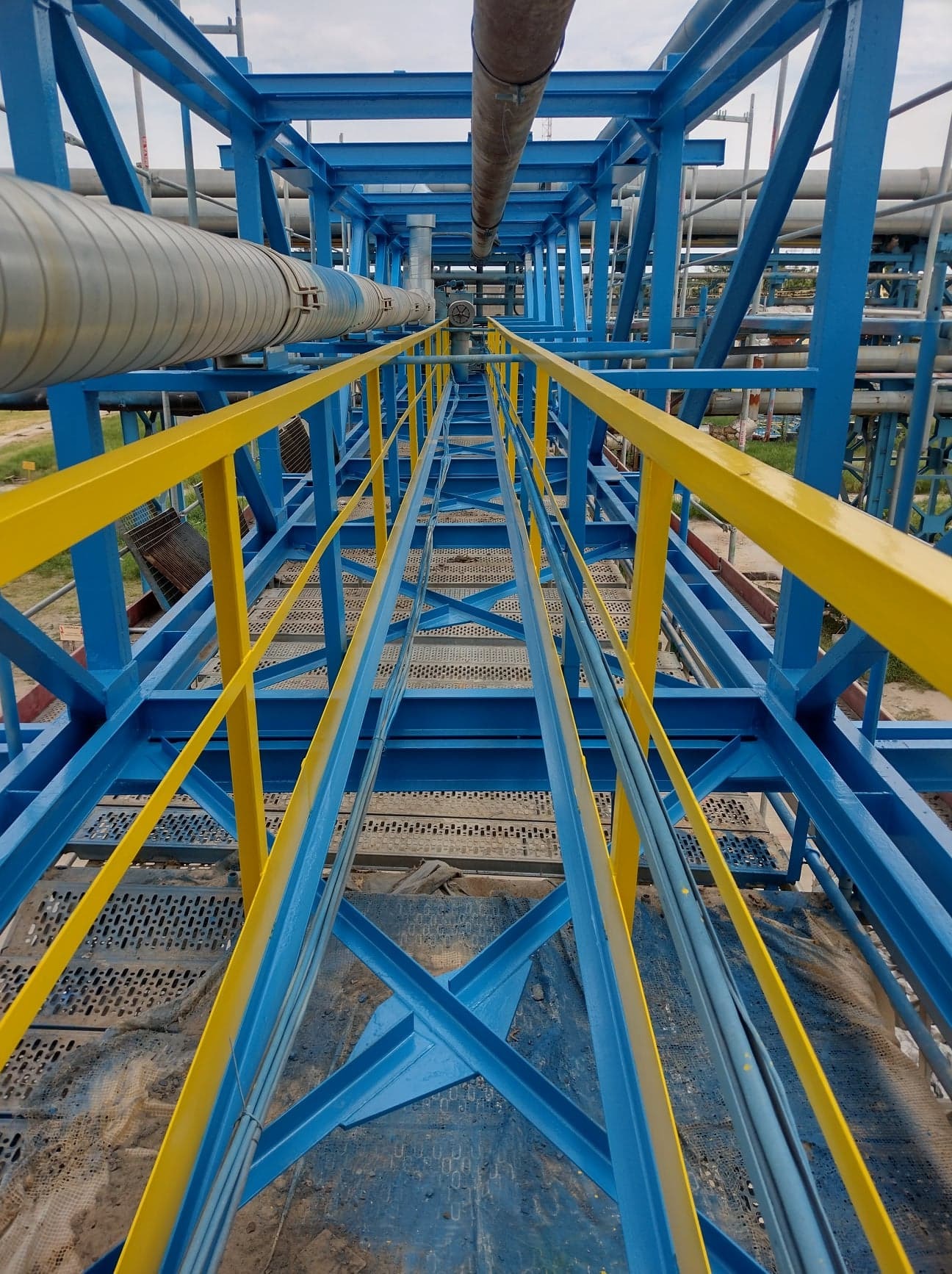Wysoka konstrukcja stalowa pomalowana na niebiesko z żółtymi poręczami, widok z perspektywy osoby stojącej na metalowej platformie, rurociągi w tle.