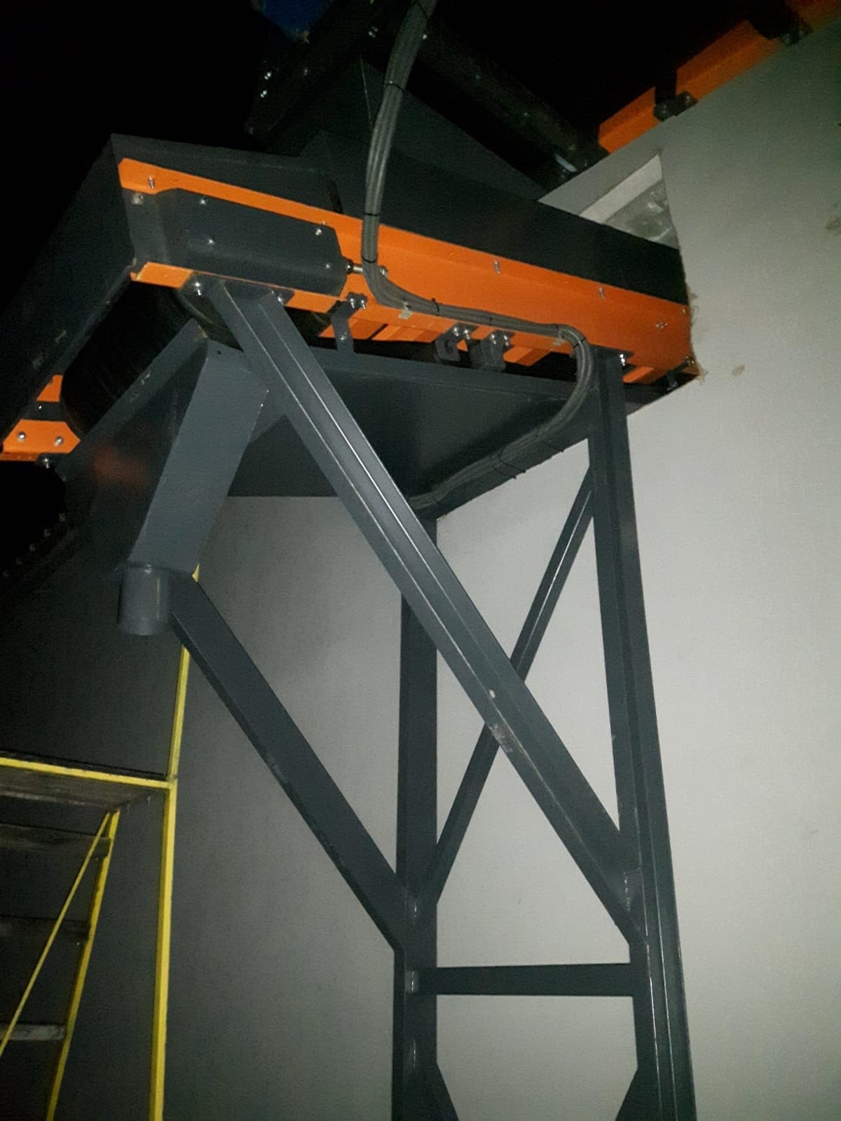 Fragment konstrukcji stalowej hali z elementami pomalowanymi na pomarańczowo, widoczne przewody i drabina malarska, oświetlenie słabe.