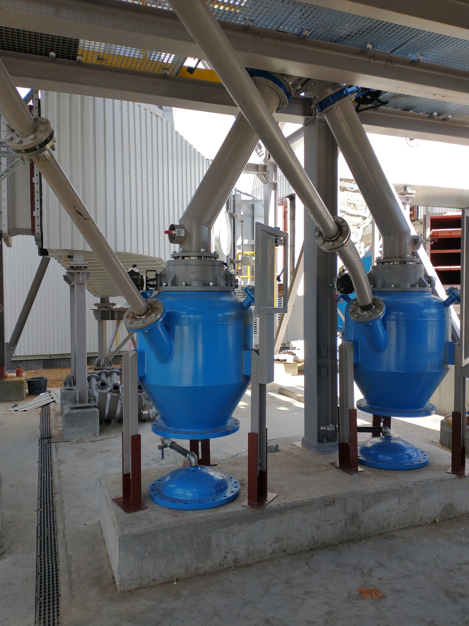 Dwa niebieskie zbiorniki przemysłowe z rurami, element instalacji w hali, wsparte na metalowych konstrukcjach, widoczne połączenia śrubowe.