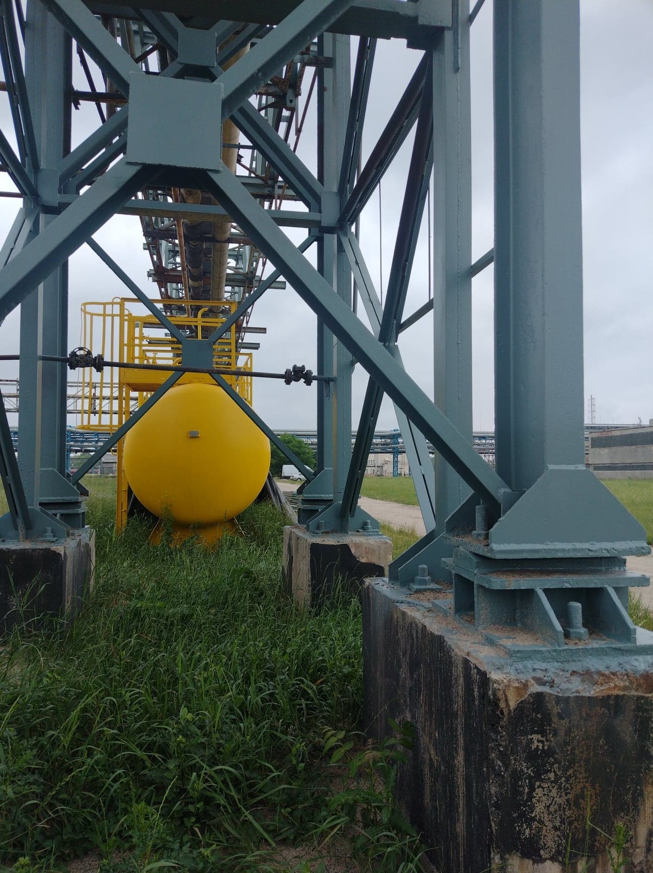 Konstrukcja stalowa podtrzymująca rurociągi, pomalowana na szaro-zielono, z żółtym zbiornikiem w dolnej części, osadzona na betonowych fundamentach porośniętych trawą.